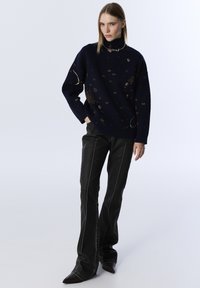 Woman wearing a black turtleneck sweater with gold star and moon patterns, black flared pants, and pointed-toe shoes, posing with one hand in pocket.