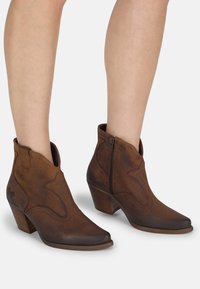 Botas de tobillo de cuero marrón con punta afilada, cremallera lateral y costuras decorativas. El tacón apilado añade altura, con un sutil logotipo.