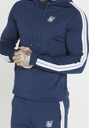 Uomo che indossa una tuta blu navy con strisce bianche e loghi sulle maniche e sui pantaloni, mani incrociate che mostrano tatuaggi e anelli.