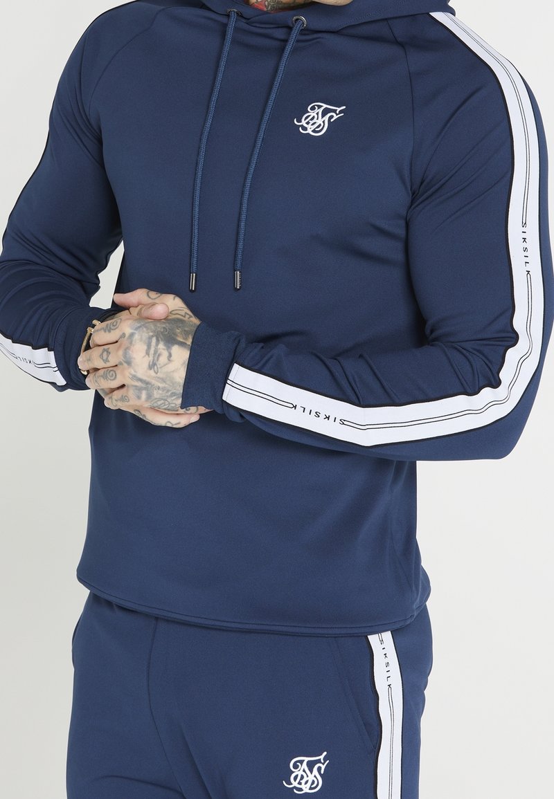 Man wearing navy blue tracksuit with white stripes and logos on sleeves and pants, hands clasped showing hand tattoos and rings.