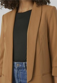 Brązowa, dopasowana marynarka z gładkiej tkaniny, z zapięciem na jeden guzik i lekkim wypełnieniem na ramionach, zestawiona z czarnym topem i dżinsami.