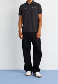 Polo shirt gris foncé avec un col zippé, arborant les logos Puma et Mercedes. Porté avec un pantalon noir et des baskets blanches.