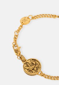 Gold chain bracelet featuring a round medallion with a detailed design. Textured links are visible, alongside a secure clasp and logo tag.