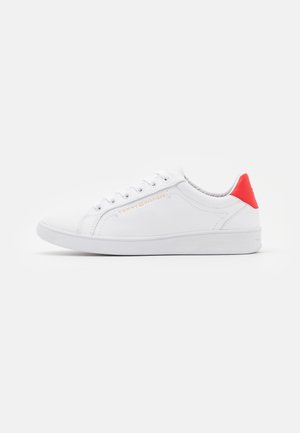 Zapatilla baja blanca con cordones blancos, pestaña del talón roja, logo dorado de Tommy Hilfiger en el lado y suela blanca sobre un fondo blanco.