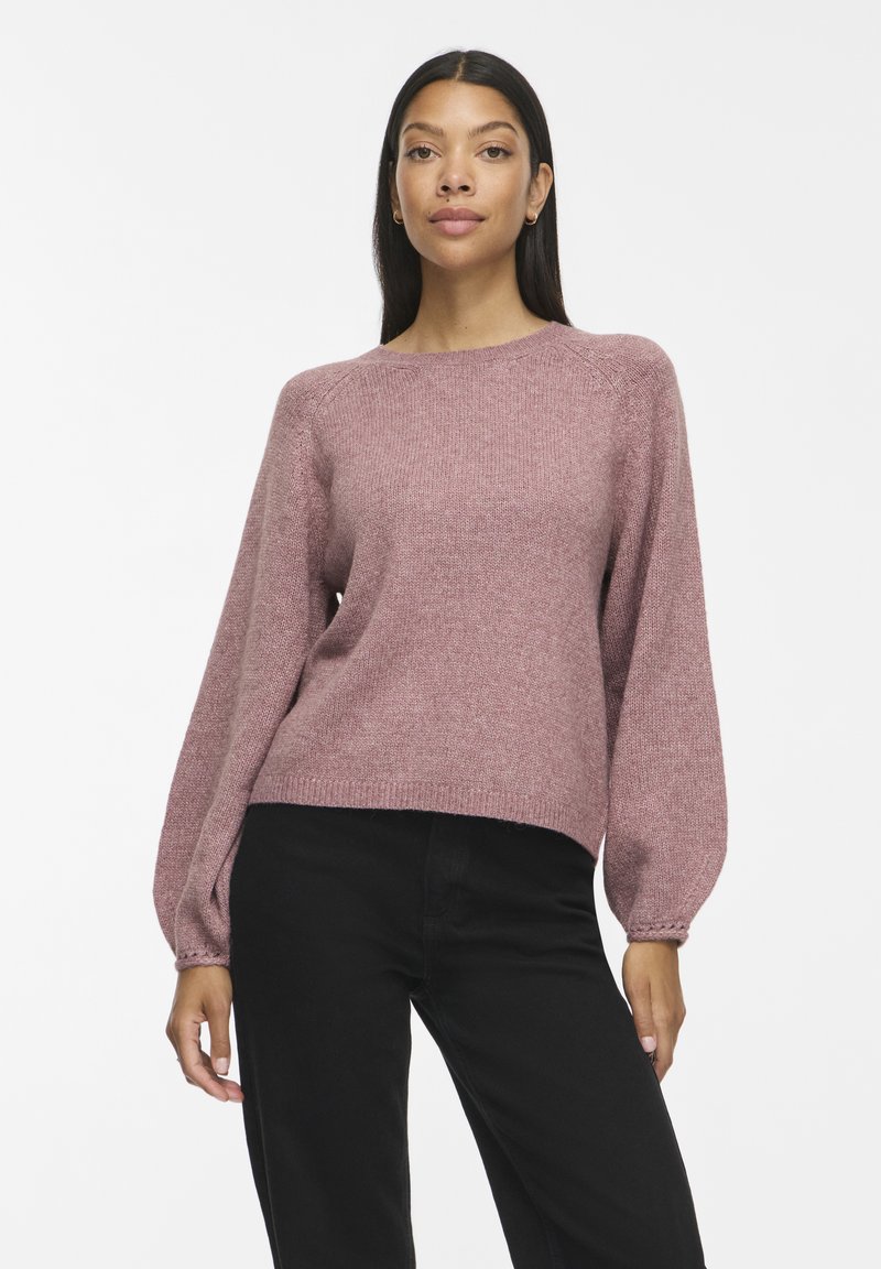 Femme portant un pull en maille mauve avec des manches ballon et un pantalon noir, debout devant un fond blanc uni.