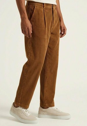 Muž v hnedých kordových nohaviciach a béžových teniskách stojaci s uvoľnenými rukami pri tele na jednoduchom pozadí.