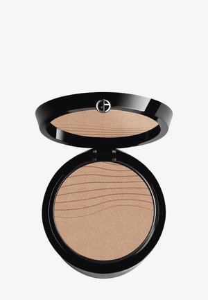 Pudder bronzer i en rund svart kompaktboks. Glatt, skimrende lys brun innside med bølge-lignende tekstur. Har en logo på lokket.