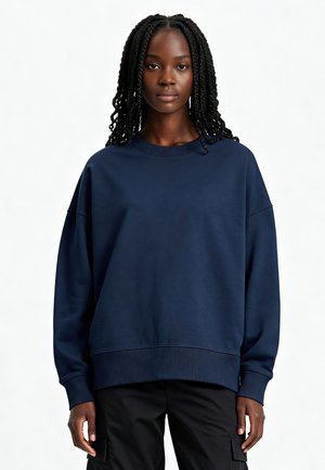 Jonge vrouw met lange vlechten, gekleed in een losse marineblauwe trui en zwarte cargobroek, staand tegen een effen lichte achtergrond.