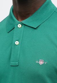 Polo vert en tissu texturé avec col classique et deux boutons. Comprend un petit logo brodé sur la poitrine.