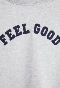 Tissu gris clair avec un texte noir brodé en arc de cercle indiquant "FEEL GOOD" sur la surface.