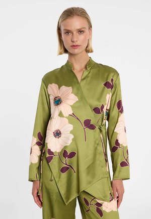 Frau trägt olivgrünes Wickeloberteil und Hose mit großem cremefarbenem und lila Blumenmuster, steht vor schlichtem weißem Hintergrund.