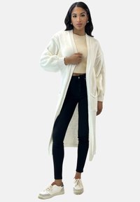 Lungo cardigan bianco con polsini a costine e tasche, indossato su un top corto beige e jeans skinny neri. Le sneaker presentano dettagli bianchi e tortora.