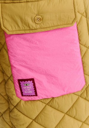 Kurtka pikowana w musztardowym kolorze, z wyraźną różową kieszenią na naklejkę w kolorze fioletowym, z napisem "Najlepsze dopiero przed nami."