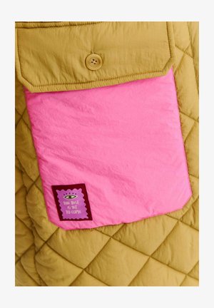 Kurtka pikowana w musztardowym kolorze, z wyraźną różową kieszenią na naklejkę w kolorze fioletowym, z napisem "Najlepsze dopiero przed nami."