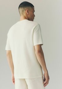Short-sleeve, cream-colored shirt made from textured fabric, featuring a simple crew neckline and a relaxed fit.