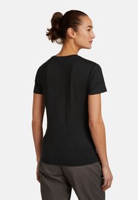 Donna con capelli castani raccolti in una coda di cavallo, che indossa una semplice maglietta nera a maniche corte e pantaloni grigi, vista di spalle, su uno sfondo bianco.