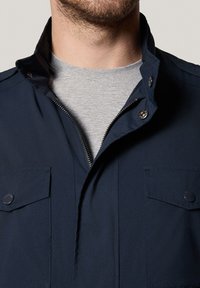 Granatowa kurtka z wysokim kołnierzem, zamkiem z przodu i dwiema kieszeniami na piersi. Materiał wydaje się gładki, uzupełniony szarym podkoszulkiem pod spodem.