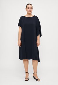 Marineblå kjole med løs, A-line silhuett og batwing ermer, laget av lett stoff; kombinert med svarte sandaletter med åpen tå.