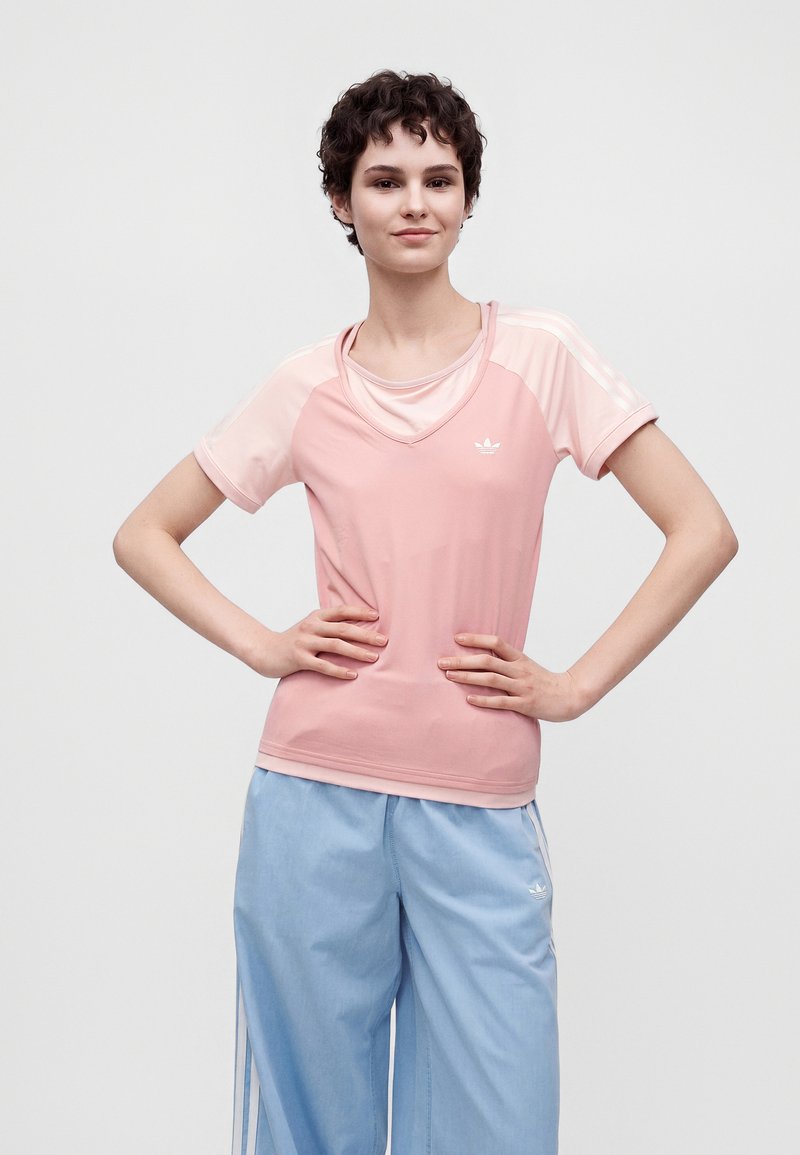 Jeune personne aux cheveux courts et bouclés portant un t-shirt rose Adidas à col en V et un pantalon de survêtement Adidas bleu clair, debout les mains sur les hanches.