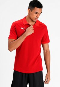 Rød polo skjorte med korte ærmer, krave og hvidt Puma-logo. Glat stof med tætsiddende design, parret med sorte shorts.