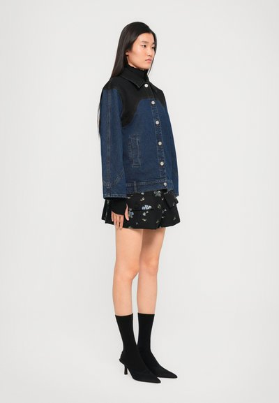 Traper jakna s crnim kožnim detaljima, tamnoplave boje, s dugmadi sprijeda, u kombinaciji s crnom cvjetnom suknjom, crnim čarapama i cipelama s oštrim vrhom.