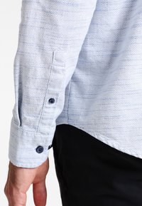Chemise bleu clair en tissu texturé. Dotée d'un poignet boutonné avec un bouton bleu foncé et de fines rayures horizontales. Coupe ajustée.