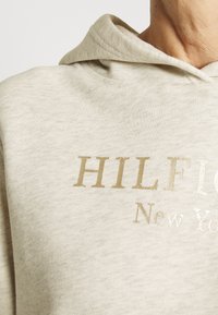 Krémová mikina s kapucí vyrobená z měkkého materiálu, s velkým zlatým a stříbrným logem, které hlásá "HILFIGER New York." Text má metalický lesk.