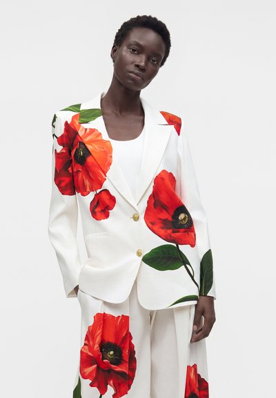 Femme portant un costume blanc avec de grandes imprimés de fleurs de coquelicot rouges sur le blazer et le pantalon, debout devant un fond blanc uni.