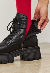 Černá kožená kotníková bota s kulatou špičkou, masivní podrážkou s vroubkováním a bočním zipem. Vnitřek je podšitý měkkým černým materiálem, který odhaluje červenou látku.