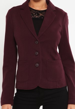Femme portant un blazer ajusté violet foncé avec trois boutons et deux poches avant, sur un top noir à encolure en dentelle.