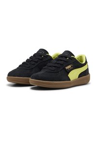 Zwarte suède sneakers met een gele PUMA-logo en accenten, met een gestructureerde bruine rubberen zool en ronde veters.