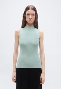 Νεαρή γυναίκα με μακριά καστανά μαλλιά, φοράει αμάνικο ανοιχτοπράσινο πλεκτό τοπ και μαύρο παντελόνι, στέκεται μπροστά σε απλό λευκό φόντο.