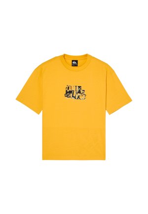 Enfant aux cheveux bouclés debout, portant un t-shirt graphique jaune, un jean ample gris foncé et des baskets noires sur fond blanc uni.