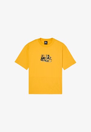 Enfant aux cheveux bouclés debout, portant un t-shirt graphique jaune, un jean ample gris foncé et des baskets noires sur fond blanc uni.