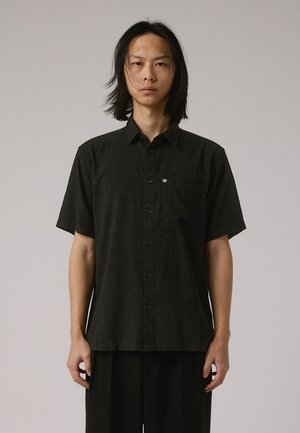 Chemise noire à manches courtes avec un motif texturé, fermeture par boutons, poche poitrine unique, et un détail logo subtil sur le côté.