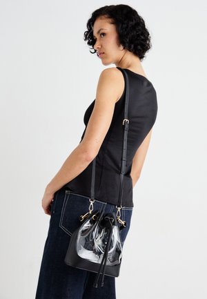 Schwarze Bucket-Tasche mit transparenten Kunststoffakzenten, goldenen Beschlägen, verstellbarem Riemen und Kordelzugverschluss, getragen über einem schwarzen Oberteil und Jeans.