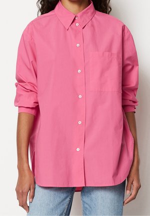 Kvinde iført en løstsiddende, knap-op lyserød skjorte med opkrogede ærmer og lyseblå jeans, stående mod en ensfarvet baggrund.