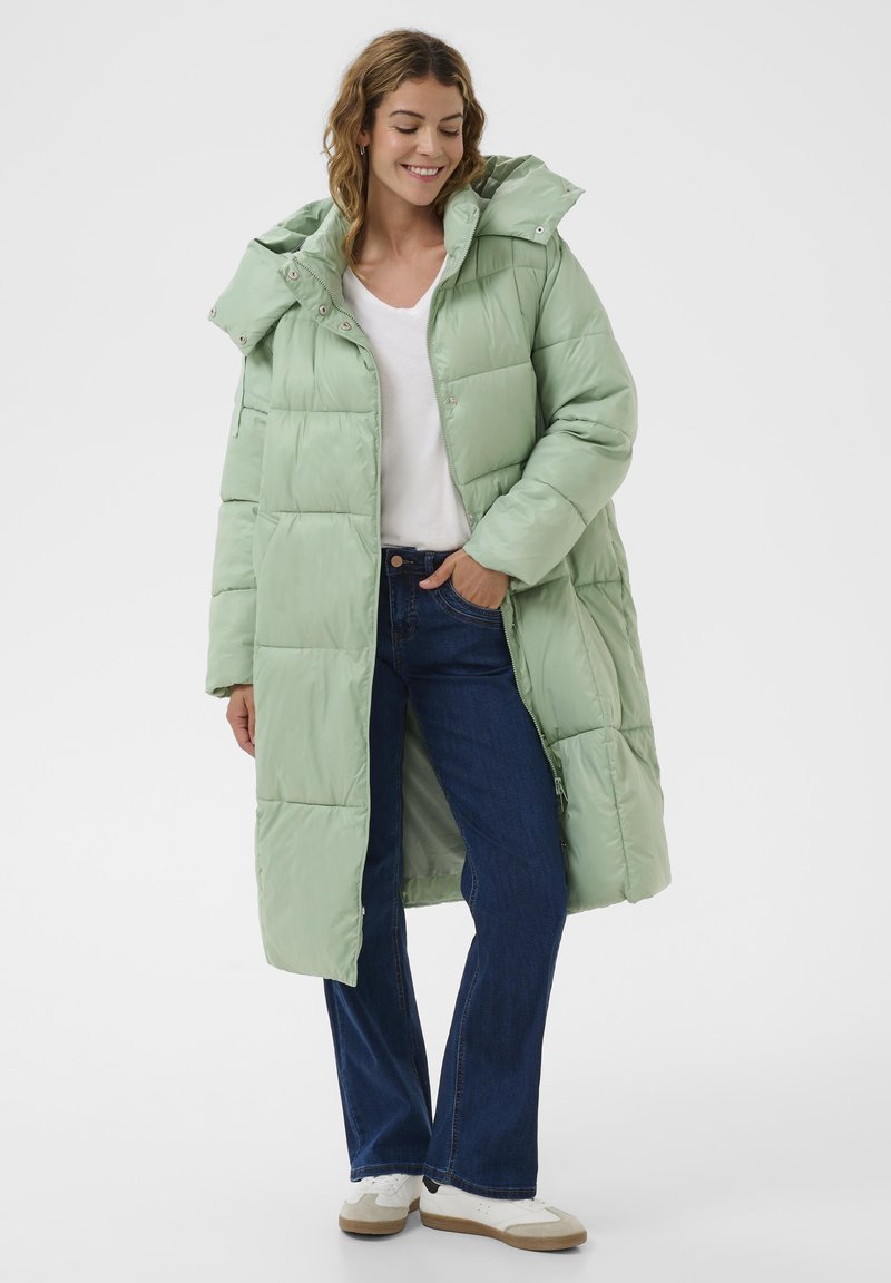 Kobieta uśmiechająca się, ubrana w długą, miętową pikowaną kurtkę, białą koszulkę, niebieskie dżinsy i białe trampki, stoi z jedną ręką w kieszeni.