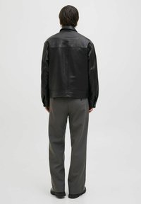 Jachetă din piele neagră cu un design minimalist, având gulerul deschis și un tiv ușor curbat. Pur tată pe pantaloni gri, evidențiind o textură fină.