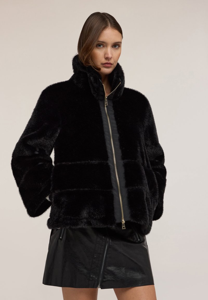 Kobieta ubrana w czarną, pluszową kurtkę futrzaną z wysokim kołnierzem i zamkiem, zestawioną z czarną skórzaną mini spódnicą na jednolitym tle.