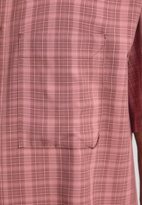 Camicia a maniche corte di colore rosa chiaro con motivo a quadretti e taschino sul petto. Il tessuto sembra leggero e strutturato, con dettagli di cucitura sottili.