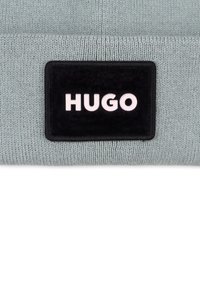 Light gray knit beanie with a black rectangular patch featuring "HUGO" in white bold font. Soft texture and fitted design for comfort.