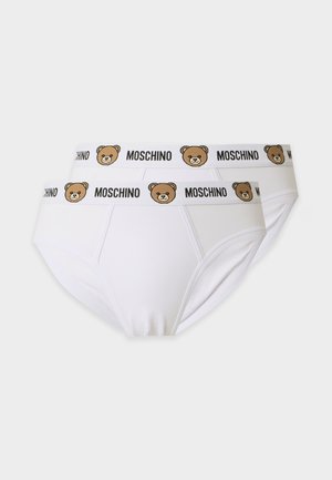Jeune homme debout, pieds nus, portant un slip blanc Moschino avec un logo ourson sur la ceinture, sur un fond uni.