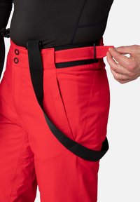 Röda skidbyxor med ett svart justerbart bälte och kardborrestängning, med sidofickor och en strukturerad tyg. Synliga knappdetaljer.