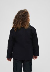 Veste noire avec une coupe droite, dotée d'un col ajustable et de manches longues. Le matériau est un tissu texturé, adapté pour le superposition.