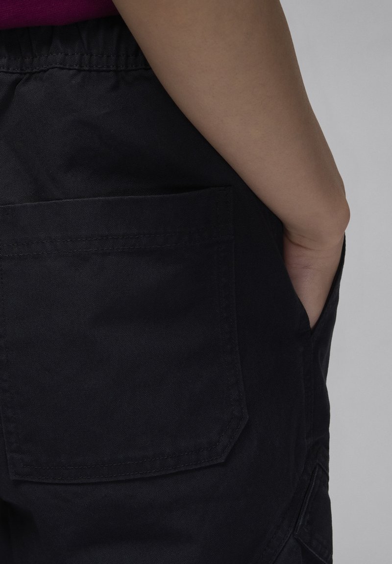 Pantalones cortos de tela oscura con una superficie texturizada, que cuentan con un gran bolsillo lateral y una cintura elástica, mostrados de lado, con una mano en el bolsillo.