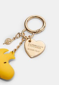 Keltainen silikooninen avaimenperä, joka on muotoiltu sarjakuvahahmoksi, kiinnitetty kultasävyiseen metalliseen avainrenkaaseen ja sydämenmuotoiseen tagiin, johon on kaiverrettu "PEANUTS."