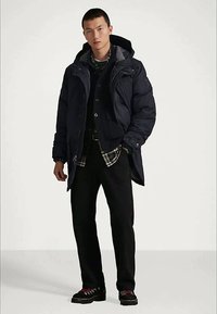 Sötétkék változatos steppelt kabát, magas gallérral, cipzárral elöl és kapucnival; egy kockás ing fölé rétegezve. Fekete nadrág és lábbeli piros részletekkel.