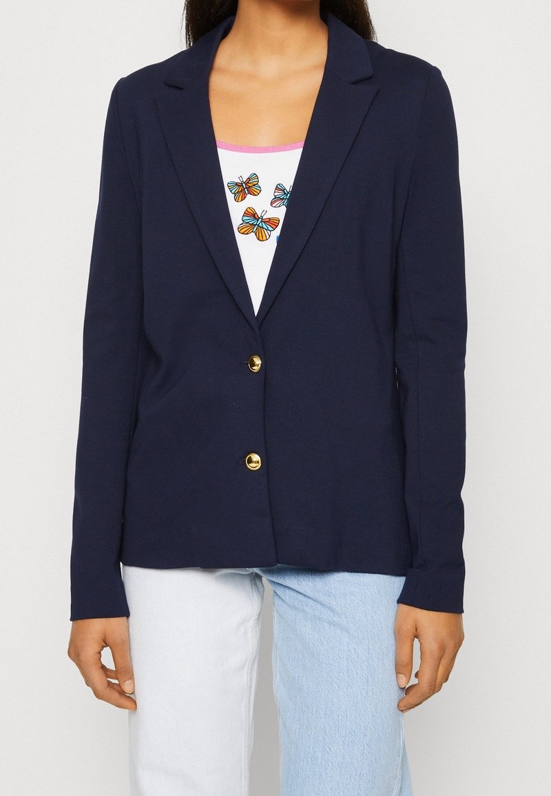 Námornícky blazer vyrobený z jemnej tkaniny, s upraveným strihom, dvoma zlatými gombíkmi a dlhými rukávmi, preložený cez biely top s pestrou vyšívkou motýlov.
