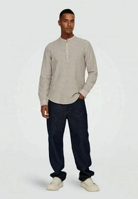 Graues Langarm-Henley-Shirt mit einem knöpfbaren Kragen, kombiniert mit dunklen Jeans und weißen Sneakers, stehend vor einem schlichten Hintergrund.